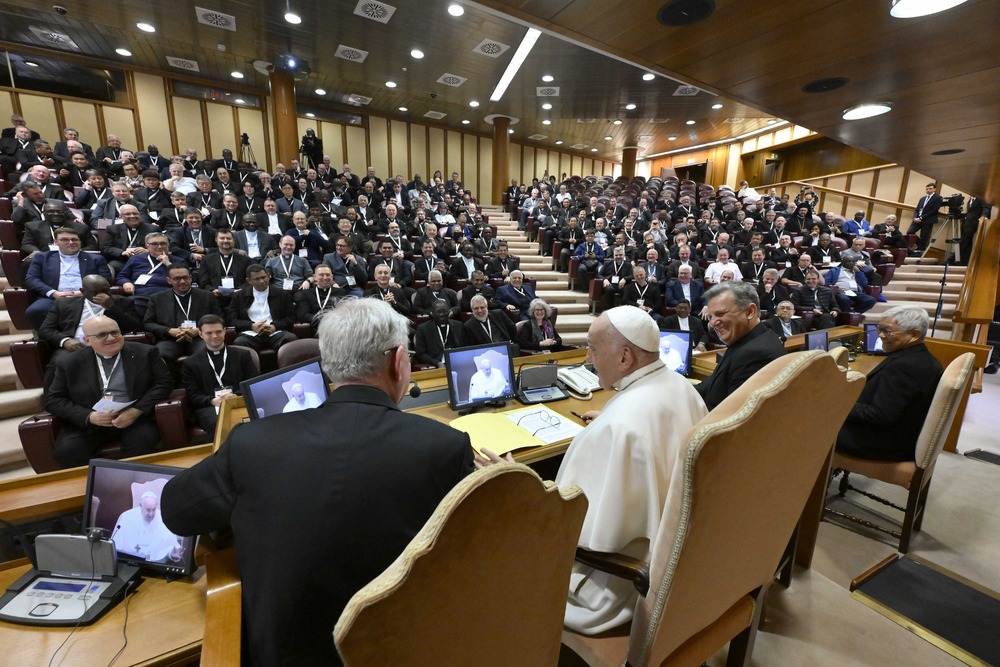 Pope pictured from behind, in front of him is audience hall filled with priests. 