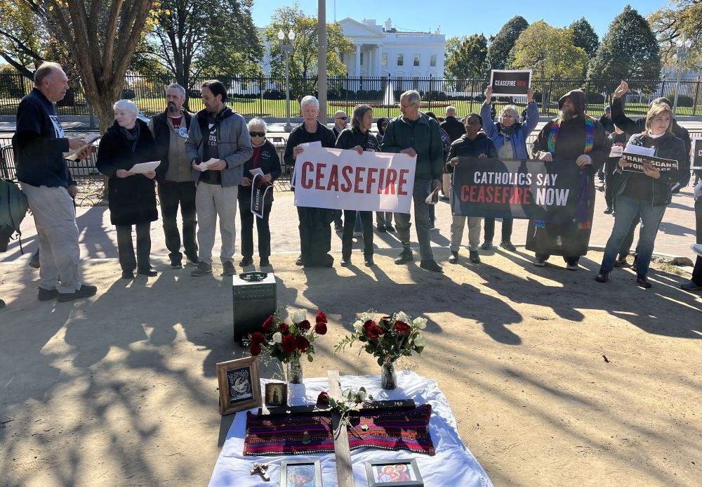 Group stands in front of a fence. White House is visible behind the fence. A person holds a sign that says, "Ceasefire."