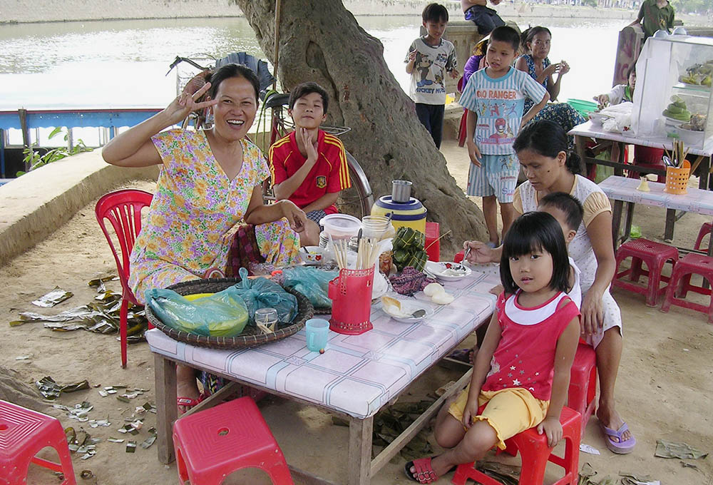 Huynh Thi Phung, quien estuvo enferma de tuberculosis, sentada desde un puesto de comida ambulante, ubicado bajo la sombra de un árbol, y rodeada de dos niños, saluda sonriente a los clientes que caminan por las aceras. 