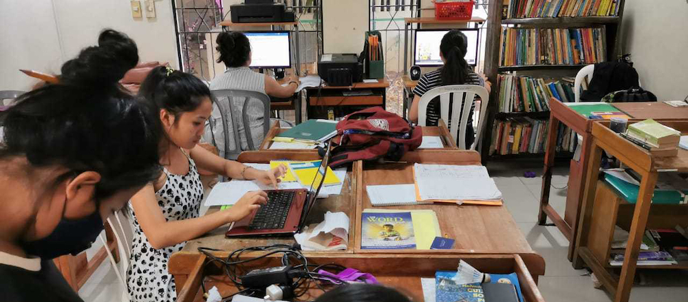 Various girls sit at tables and with computers and tablets working on school