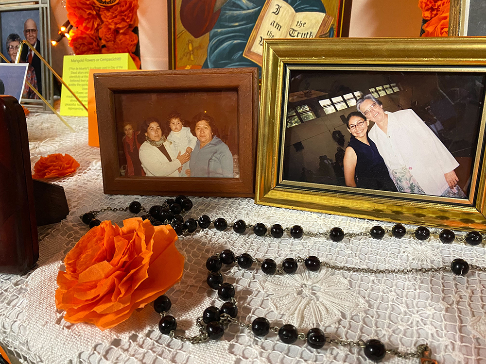 Two photographs sit on a table with a white lace tablecloth with a black rosary and orange poppy in front of them