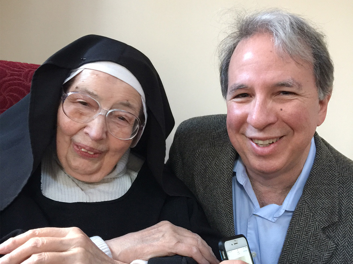 An elderly white woman in a black nun's habit and glasses poses with an older white man in a light blue shirt and gray suit jacket
