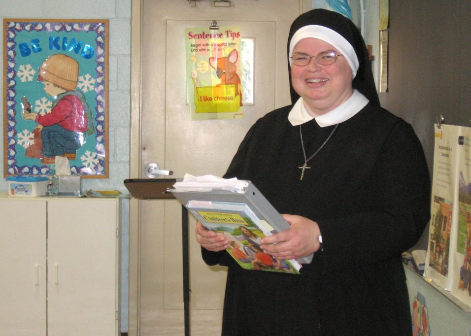 A white woman in a black nun's habit and glasses smiles at the camera, holding a gray binder and a book with both hands