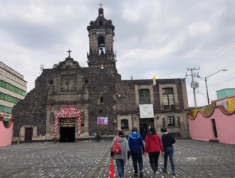 Al fondo la antigua iglesia La palmita, en donde las Hermanas Oblatas del Santísimo Redentor tienen su base de operaciones; al frente, en la plazoleta, una religiosa y otras perosnas se acercan al templo. 