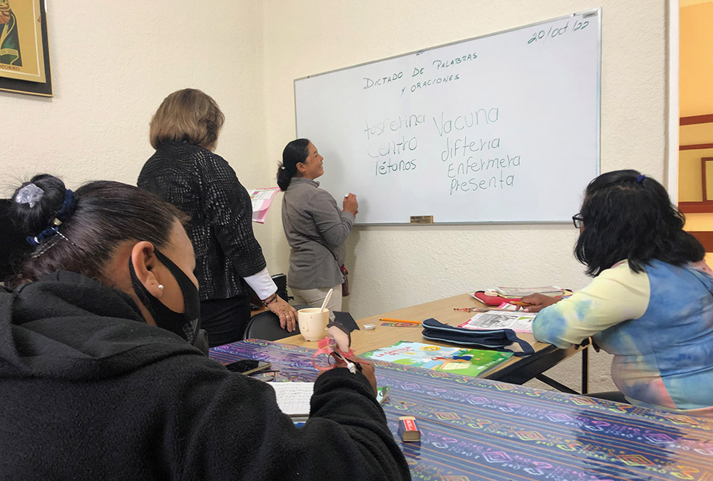 una mujer llamada Josefina practica su vocabulario en una pizarra, mientras la Hna. Rosa Aguayo, detrás de ella, supervisa, y sus compañeras la observan.