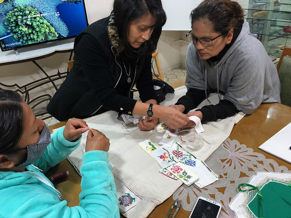 La Hna. Oblata María Rosas, en el centro y frente a una mesa, enseña patrones clásicos de bordado y técnicas de costura a dos mujeres. 