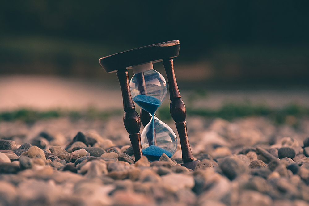 An hourglass with blue sand sits upon a field of rocks. The hourglass is half-full.