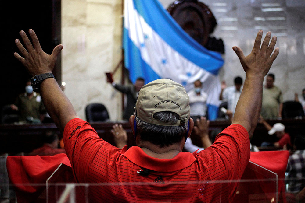 Un hombre de espaldas con camisa roja en primer plano y al fondo la bandera de Honduras ondeada en el interior del Congreso. 