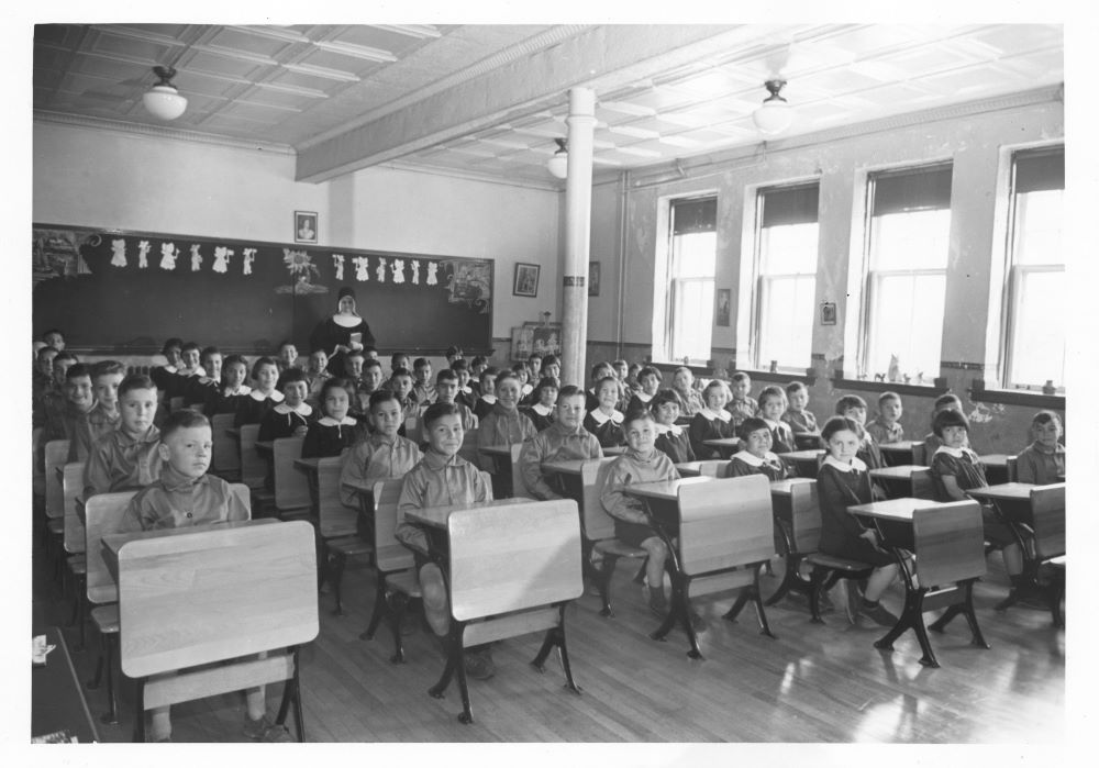 Nun stands in back of a classroom with children sitting at desks.