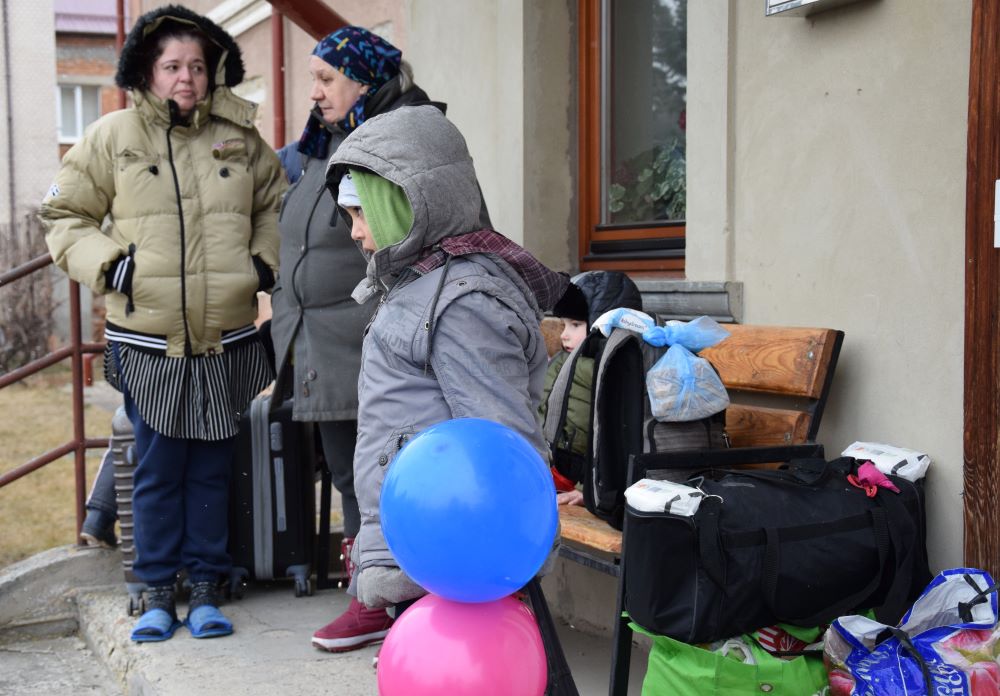 People dressed in winter coats stand on porch, amid bags and luggage. 