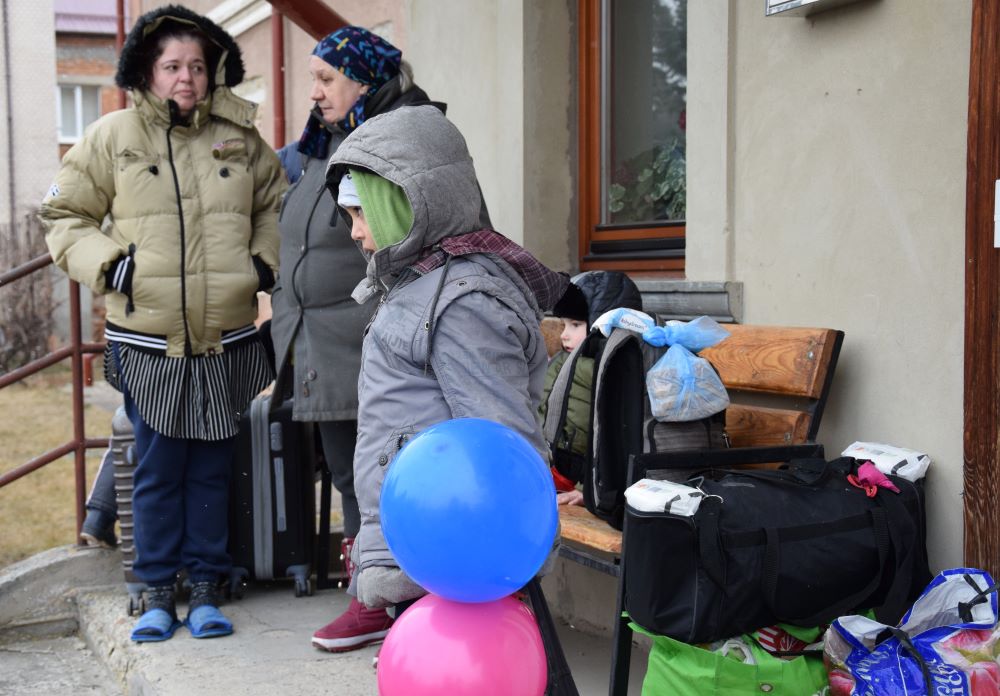 People dressed in winter coats stand on porch, amid bags and luggage.