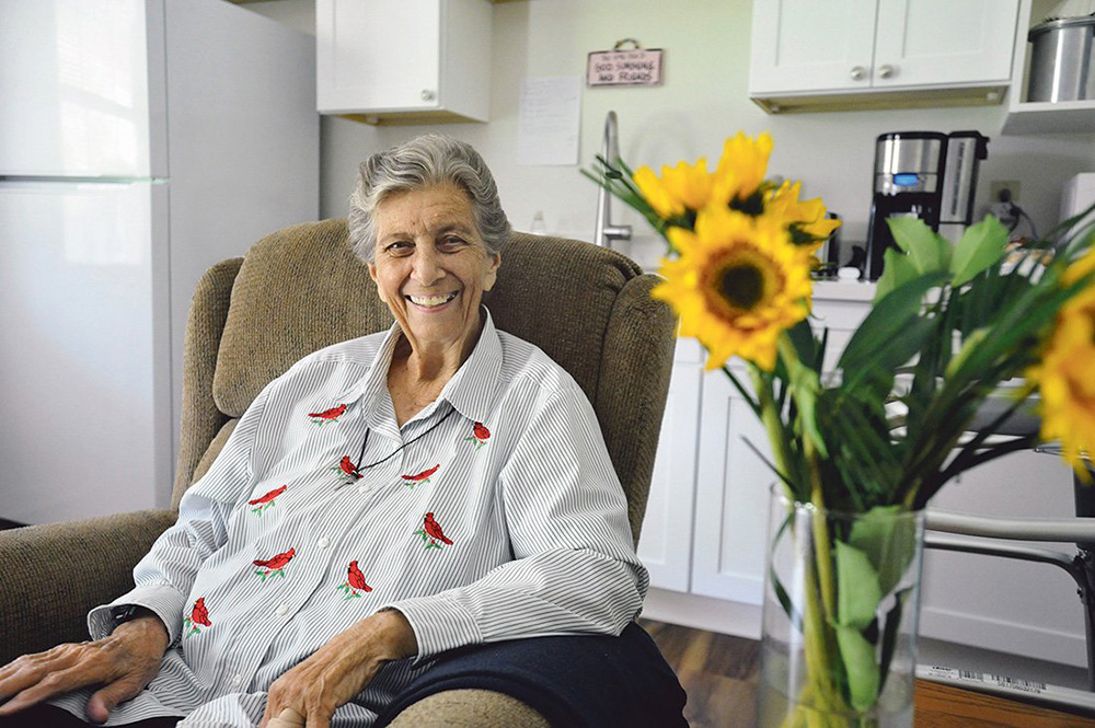 An older white woman sits in a chair smiling at the camera next to a vase of sunflowers
