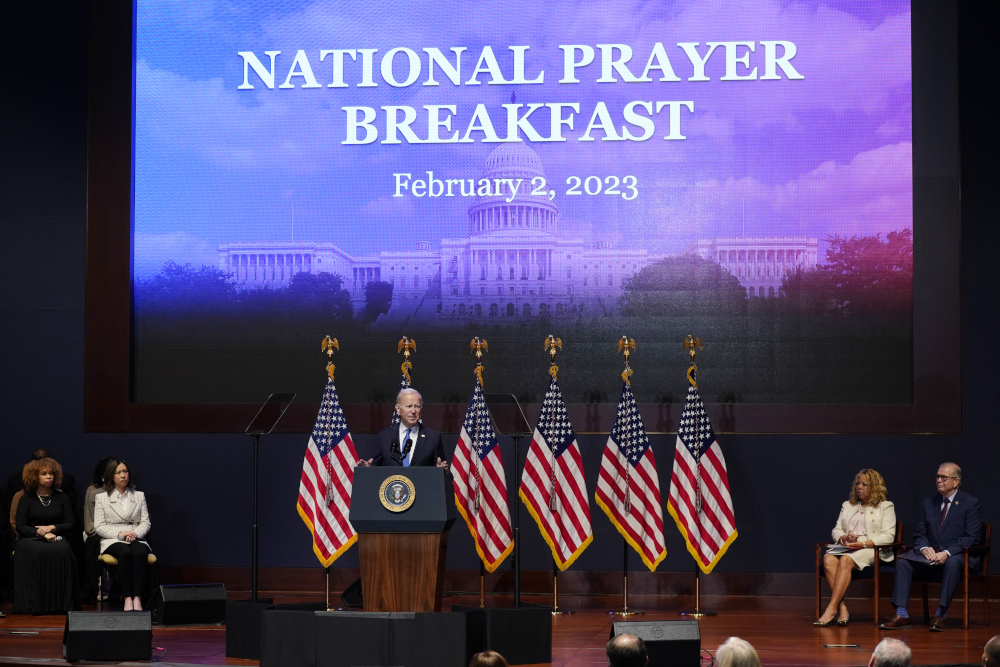 Biden stands behind a podium in front of several American flags and a projector screen that says "National Prayer Breakfast"