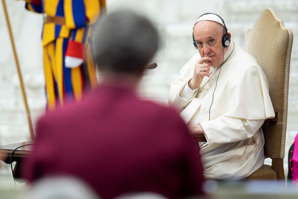 A person's back is show, as Pope Francis francis listens. He is wearing headphones. 