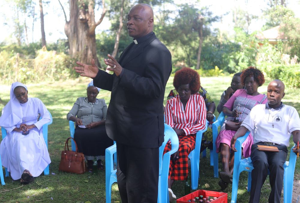 Priest stands and talks to a group of people sitting in blue chairs.