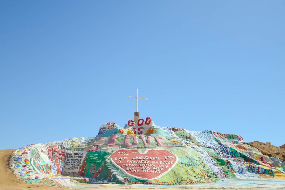 A cross stands atop a hill that is decorated with hearts and messages that say things like "God is love."