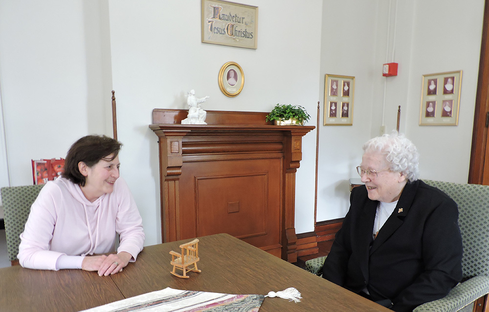 Two sisters are talking while seated and leaning on a brown table in the white visiting room of the Convent of Our Lady of the Angels in Pennsylvania.
