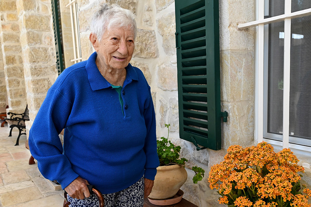 An elderly white woman in a blue long-sleeved shirt poses for a photograph next to a home with orange flowers