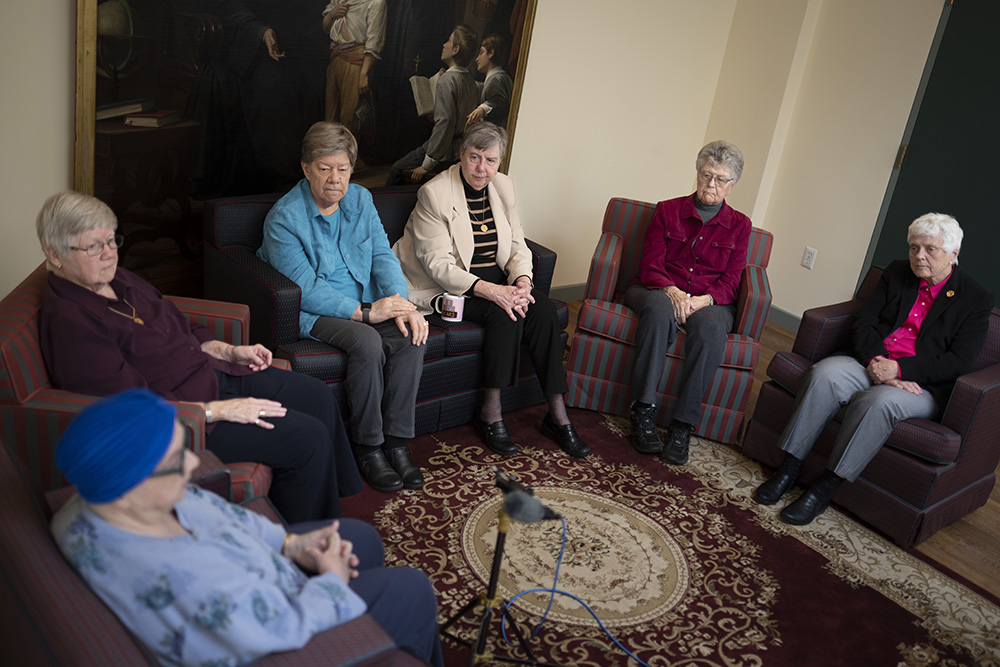 Older women sit in chairs in a circle in front of a large painting