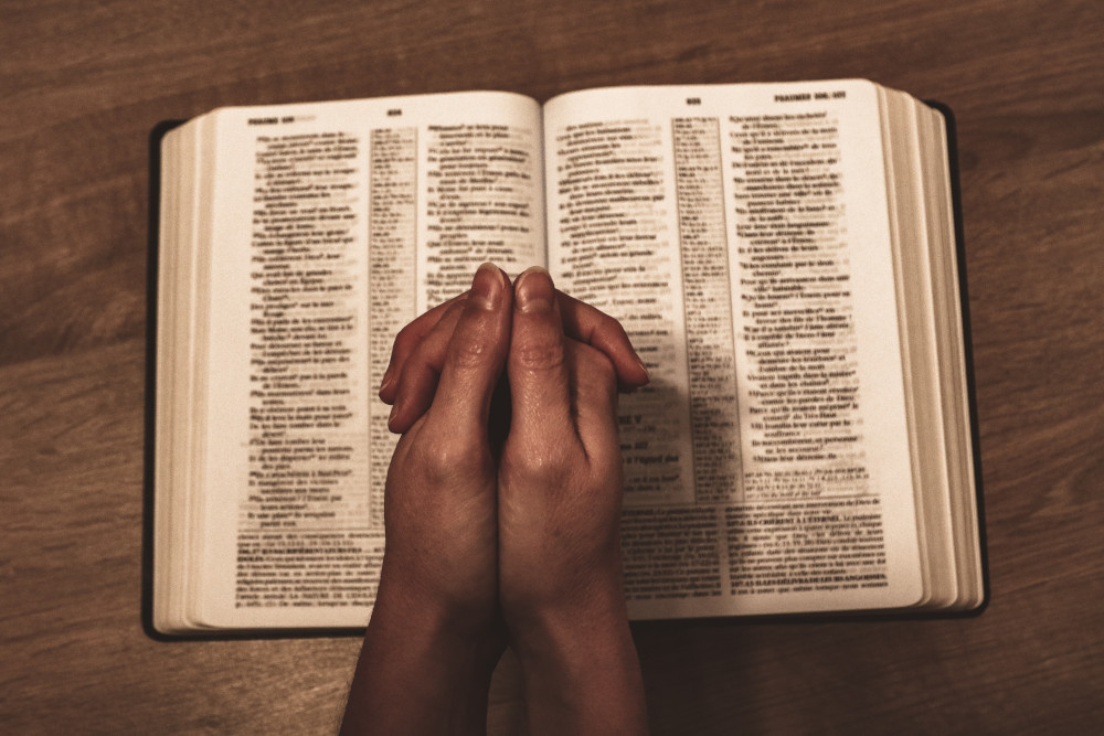 A person's hands are clasped over a bible in prayer (Unsplash/Jametlene Reskp)