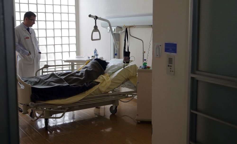 A man wearing a doctors coat talks to a patient in a hospital bed.