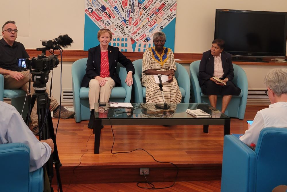 Three women sit on a couch as they talk to group and a camera records them.