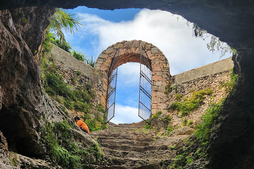 A view from a cenote/cave looks up into the sky through a gated archway in Maní, Yucatan, Mexico (Unsplash/A T Ø M E)