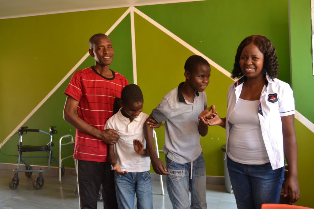 Man in red shirt supports his son as he stands next to another son and teacher. The teacher holds the other son's hand to help him stand.