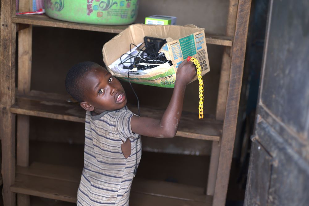 A boy looks at the camera as he lifts a battered box from a shelf. 