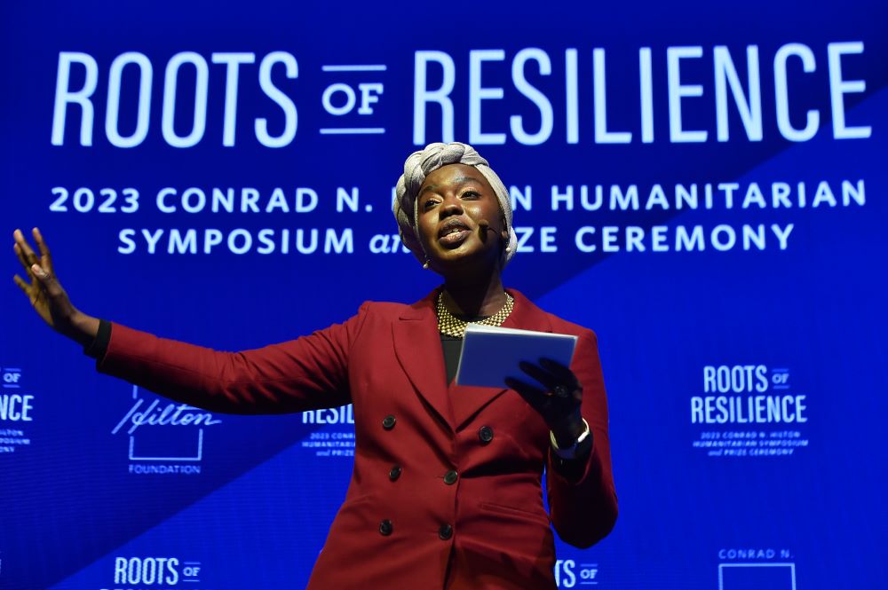 Woman holds papers and gestures with one arm. Behind her is written, "Roots of Reliliance."