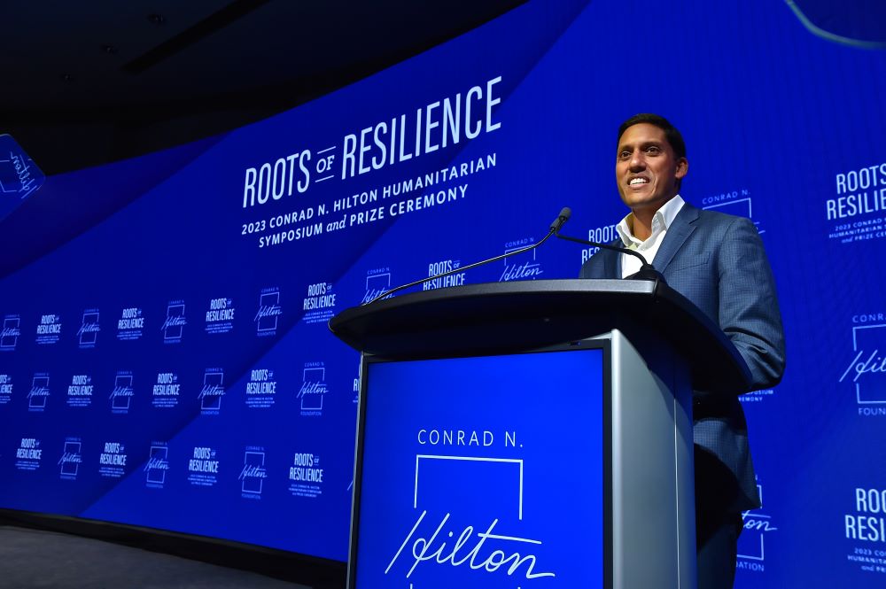 man stands at podium behind microphone. Behind him is written "Roots of Resilience."
