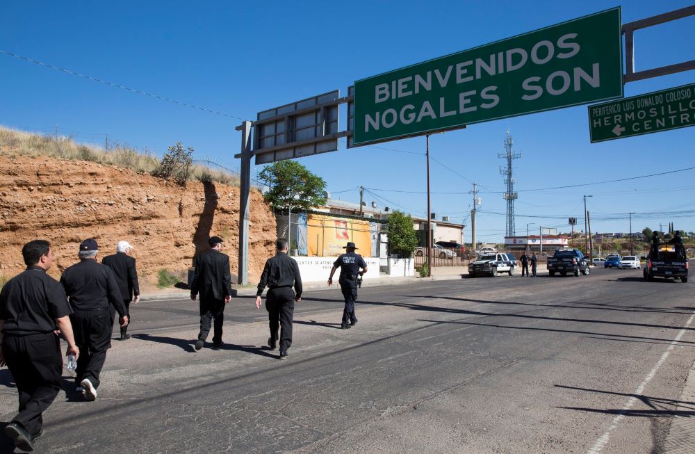 People walk on road with sign ahead: "Bienvenidos Nogalos Son"