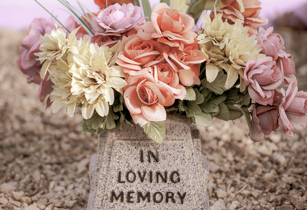 A photo illustration displays a memorial flower arrangement, in a container bearing the words "In loving memory" (Unsplash/Sandy Millar)