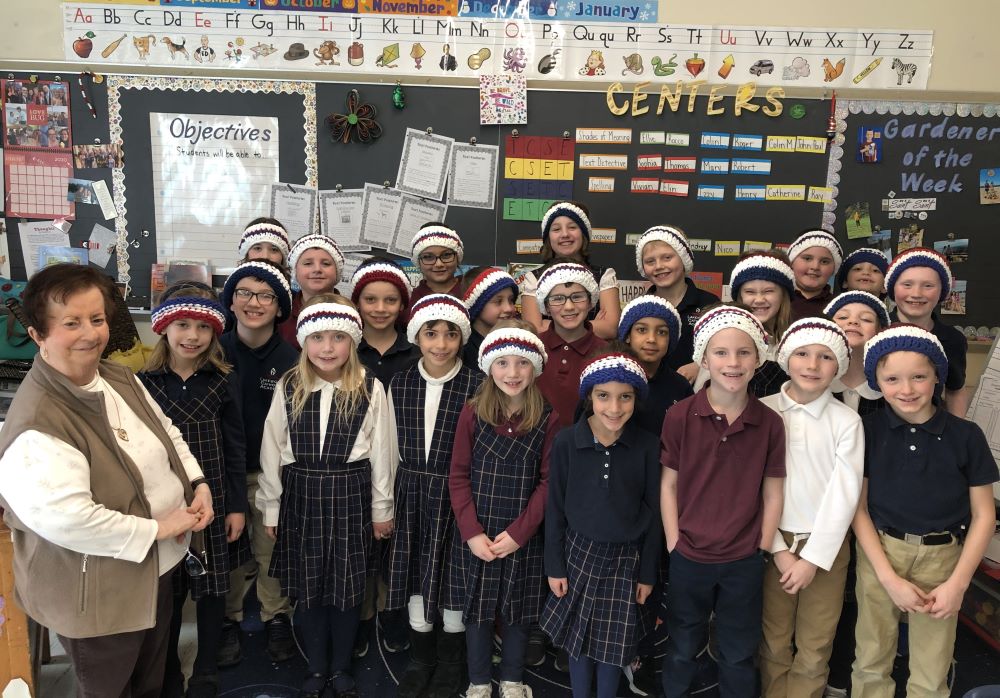 Woman stands with a group of children in a classroom. The children are wearing crocheted headbands,