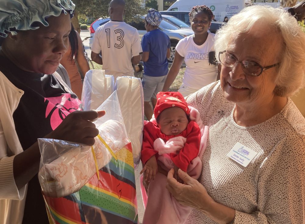 A woman stands with another woman, holding a baby.