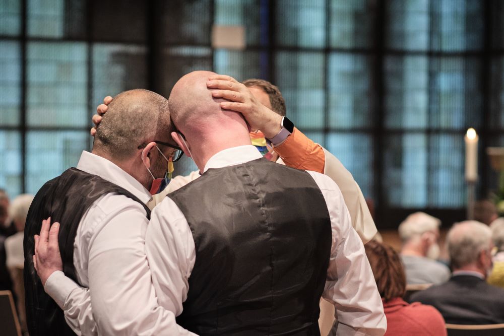 Two men with arms around each pther are seen as a priest puts his hand on nne of their heads in a blessing.