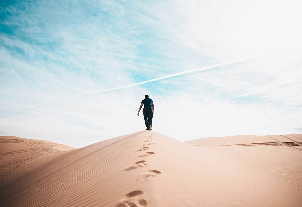 A A person walks up a hill of sand in a desert setting, with a blue sky in the background, and footsteps behind the person walking.(Unsplash/Nathan McBride)