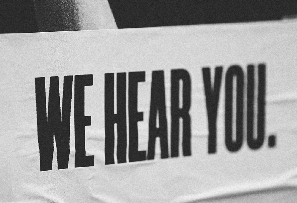 A black-and-white image features a sign with all-caps text that reads: "We hear you." (Unsplash/Jon Tyson)
