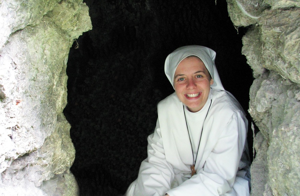 Sister Clare Crockett, 2011. The sister wears white habit, and crouches in grotto, smiling broadly.