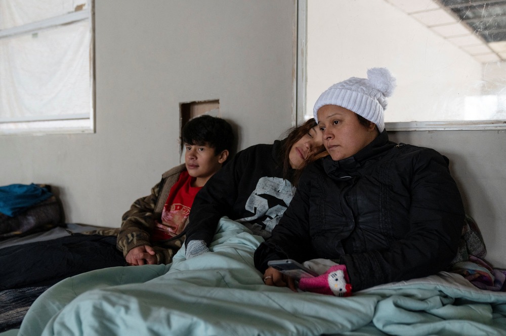 Denise Mendez, a Honduran migrant, and her children, Piedras Negras, Mexico, Jan. 20, 2024. Mendez and children wear winter clothing and sit together beneath blanket.