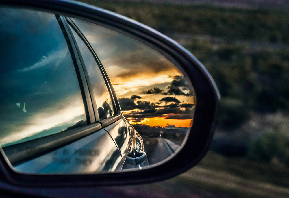 A car's rearview mirror shows a horizon full of bright orange and dark clouds. (Unsplash/Jake Weirick)