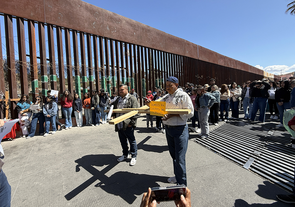 A participant in a symbolic Via Crucis held Saturday in Nogales, Mexico, to honor migrants uses a smartphone (at bottom) to take a picture of the group that includes a man holding a wooden cross next to the border wall with Arizona. The orange sign reads, "We are the body of Christ." (Anita Snow)