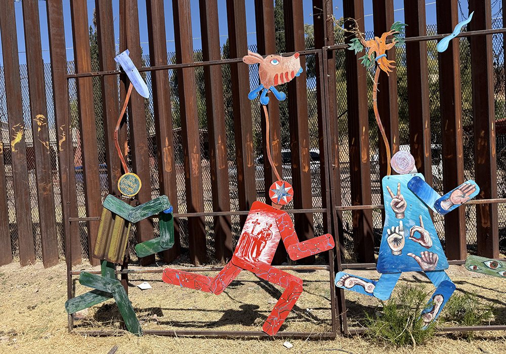 Participants in a symbolic Via Crucis that Kino Border Initiative organized April 5 in Nogales, Mexico, passed through the street alongside the border wall of tall steel bollards that often features pro-migrant art, including this one featuring a Border Patrol figure chasing two people. (Anita Snow)