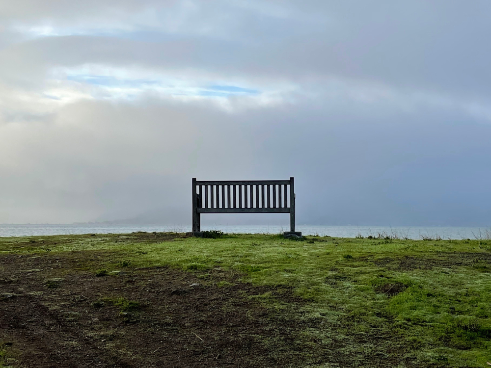 Bench A lone bench on patchy grass overlooking a cloudy sky with patches of sunshine shining through.