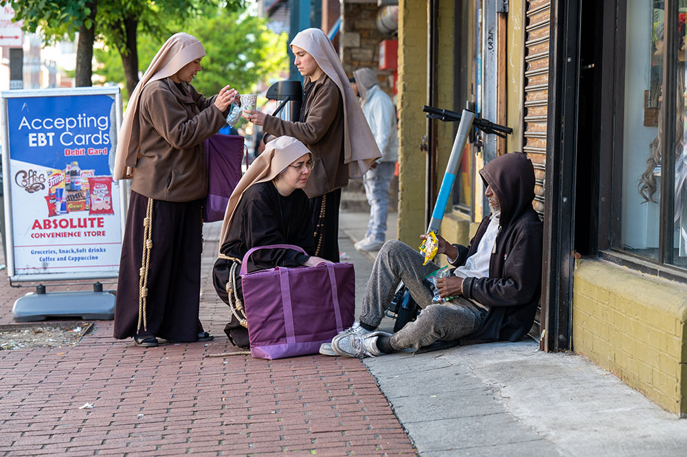 Sister Giovana of the Most Holy Name of Jesus (squatting) joins Sister Samaritan of Scourged Love (left) and Sister Maria Clara of the Crucified in giving food to a man near Lexington Market in Baltimore May 3, 2023. (OSV News/Catholic Review/Kevin J. Parks)