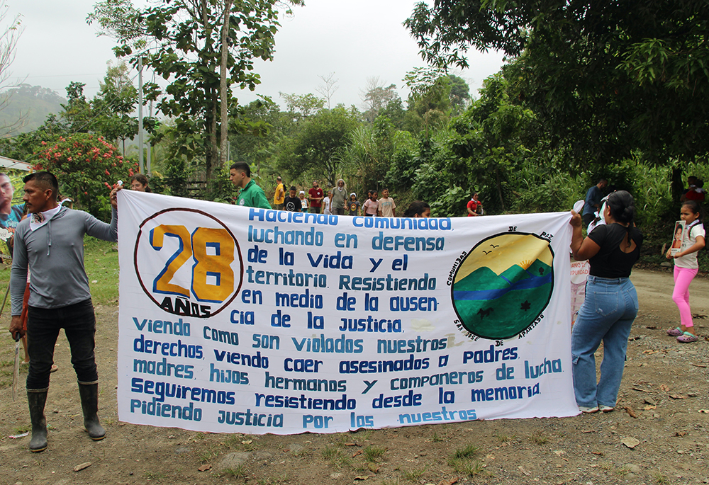 Community members hold a sign during their procession to the gravesites of members killed in 2024. It reads: "28 Years: Building a community fighting in defense of life and territory — resistance in the midst of the absence of justice, seeing how our rights are violated, seeing fathers, mothers, children, brothers and sisters and companions in the struggle fall assassinated, we continue resisting from memory, demanding justice for our loved ones." (Tracy L. Barnett)