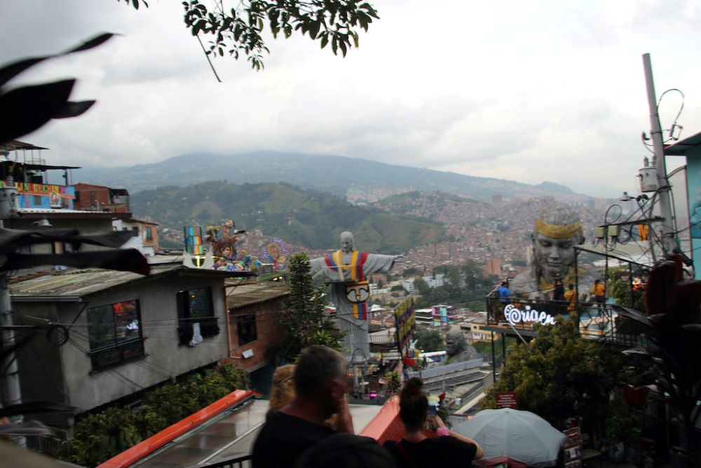 View includes flashing neon and tin-roofed homes stacked up the hillside. A tricolor-draped Christ, a massive gorilla and a concrete woman's head, an icon representing a local craft beer called “Origen,”overlook the scene. View includes flashing neon and tin-roofed homes stacked up the hillside. A tricolor-draped Christ, a massive gorilla and a concrete woman's head, an icon representing a local craft beer called “Origen,”overlook the scene.