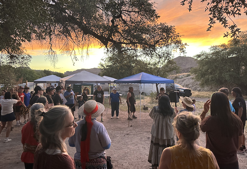 A crowd, including sisters, listening to an Apache Stronghold ally and artist rap during recent Oak Flat demonstrations. Among those who spoke at the gathering was Wendsler Nosie, an Apache elder and leader of Apache Stronghold. Nosie told the visitors of the need "to stand together" and find common ground among different groups. (Courtesy of Land Justice Features)