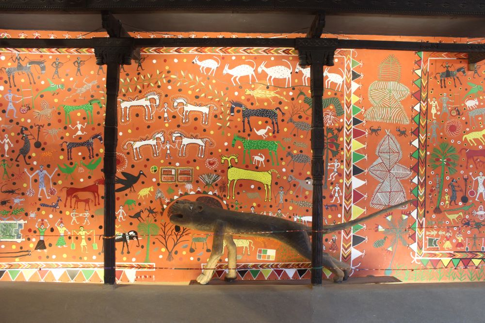 A vibrant Warli-style mural features tribal art, animals and human figures, with a life-sized animal sculpture in the foreground. This artwork represents the deep connection of Indigenous communities with nature and their cultural heritage. 