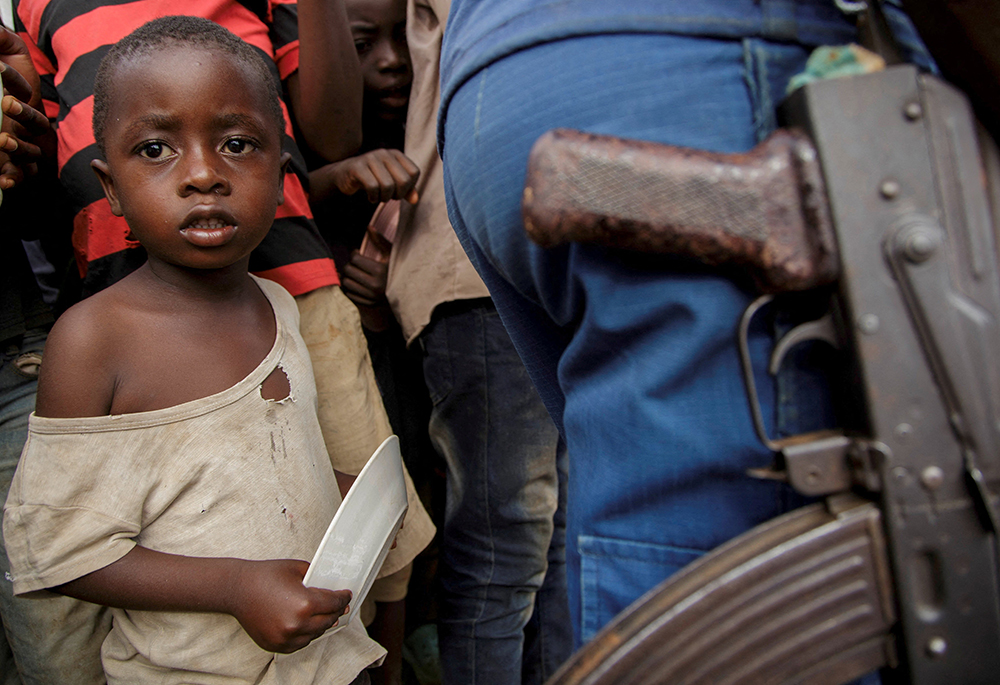 A child stands next to a police officer as displaced families wait for food at Rugombo Stadium in Burundi Feb. 18, 2025, after Congolese fled from renewed clashes between M23 rebels and the armed forces of the Democratic Republic of the Congo (FARDC). (OSV News/Reuters/Evrard Ngendakumana)